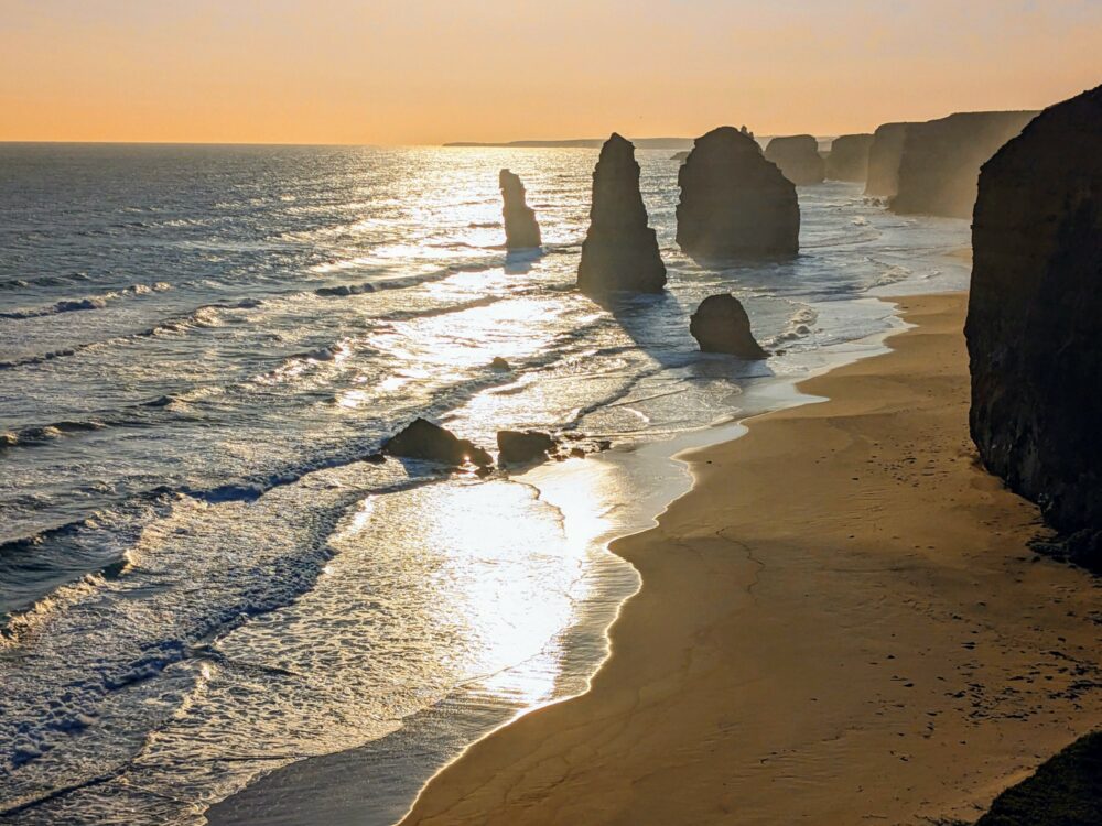 Sunset at the 12 Apostles on the Great Ocean Road. Several limestone rock formations just offshore in the ocean, with an orange sunset glow in the background.