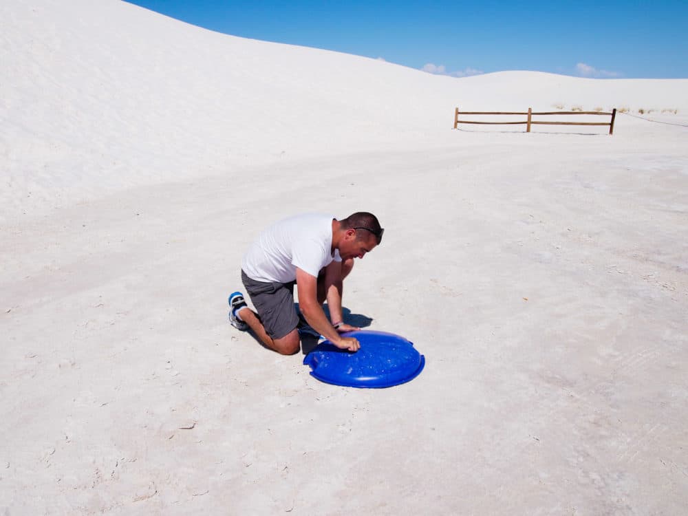 Man kneeling down at the base of a white sand dune, applying wax to a round plastic board