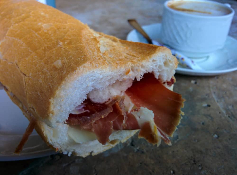 Closeup of a bread roll with Spanish ham inside, and a cup of coffee behind