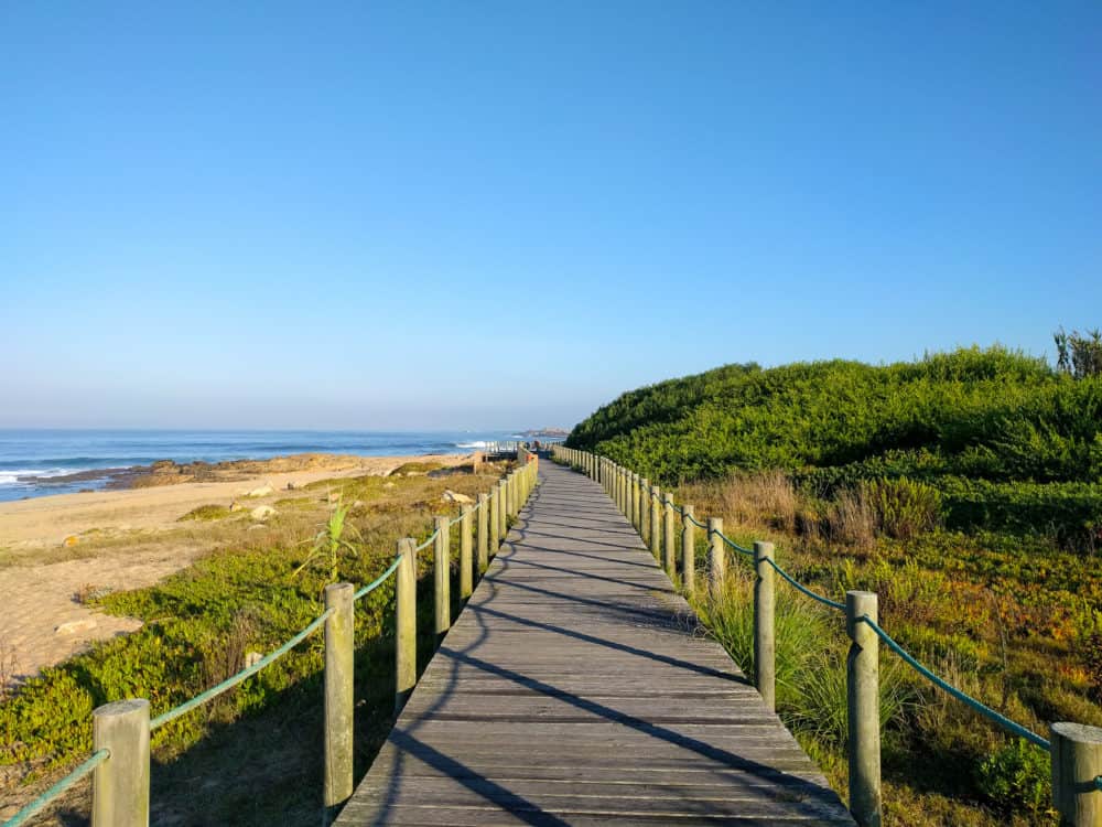 Long wooden boardwalk with a rope handrail on both sides. On the right hand is grass and a small embankment, on the left is sand and the ocean.