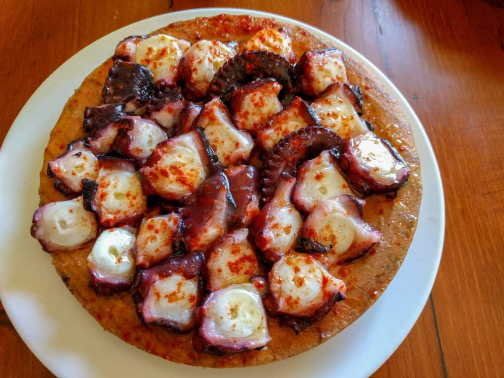 A wooden platter of grilled octopus, cut up and sprinkled with paprika and oil, sitting on a white plate on a wooden table.