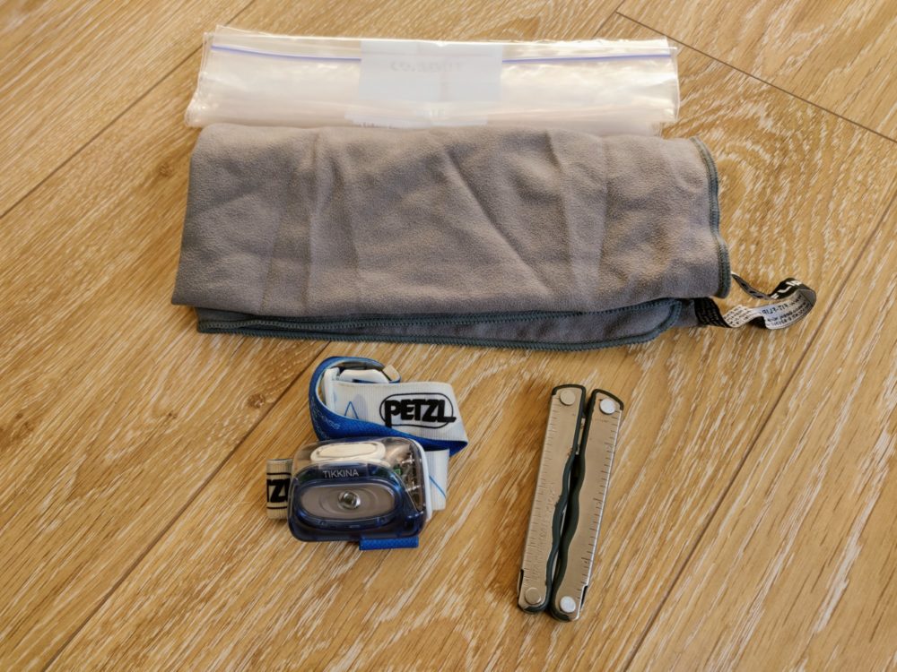 Headlamp, towel, multi-tool and plastic bags beside each other on a wooden floor.