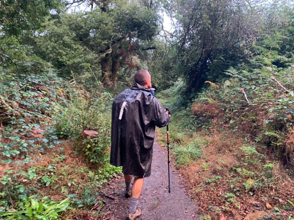 Author walking in the rain wearing a poncho on a narrow trail with trees alongside.