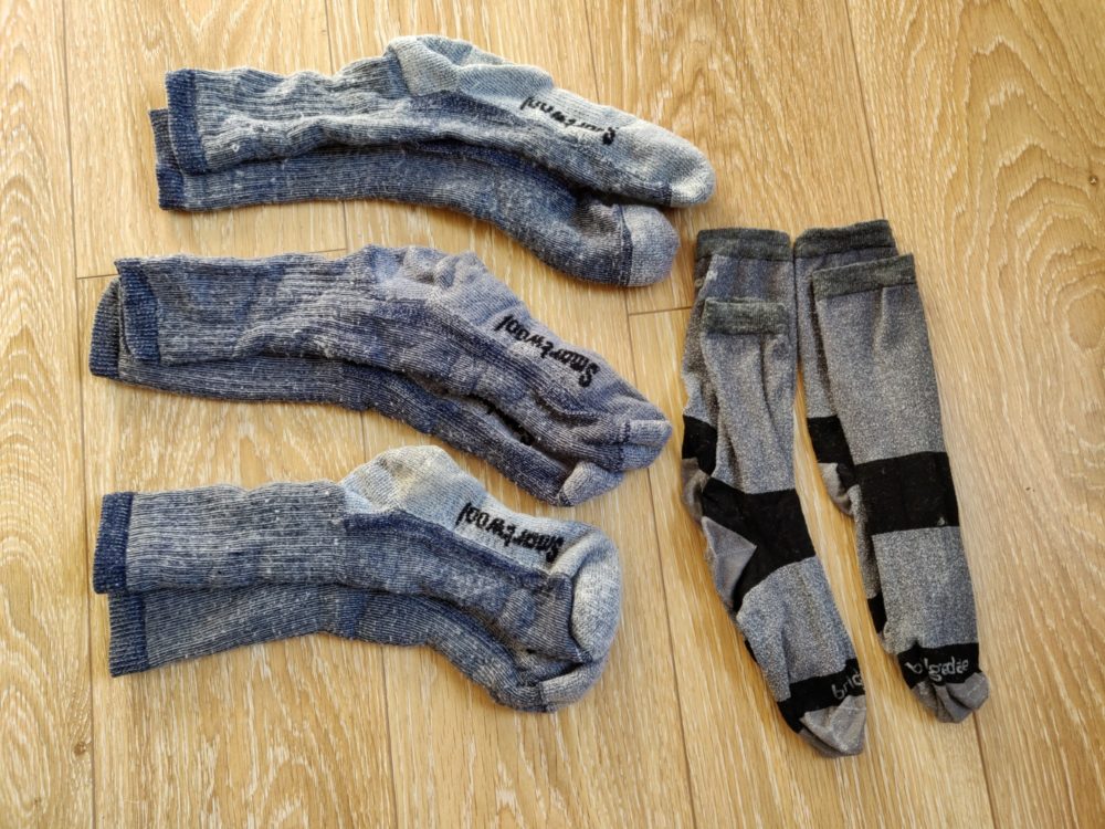 Five pairs of hiking socks: three pairs of woollen ones, two pairs of cotton liners, on a wooden floor