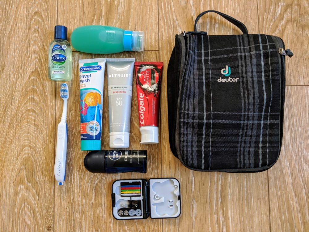 Toiletry bag, tubes of toothpaste, travel wash, and sunscreen, deodorant, sewing kit, shower gel, and toothbrush, on a wooden floor.
