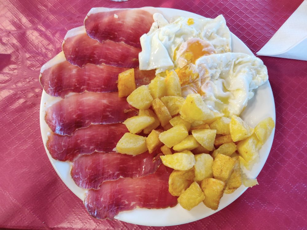Plate of food on the Camino Primitivo with thin slices of ham, fried potatoes, and egg.