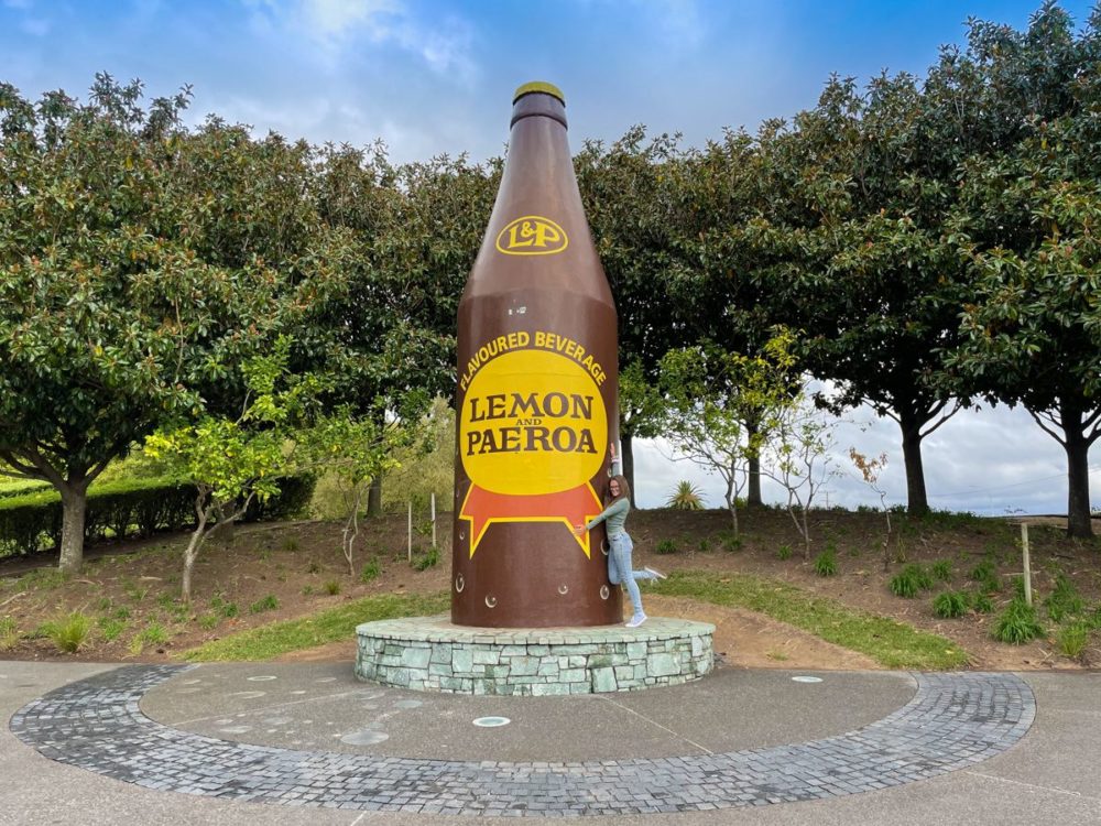 Person standing beside large sculpture of an L&P bottle in Paeroa