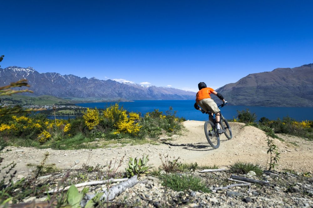 Person on mountain bike riding on a dirt trail overlooking a lake, with mountains in the background