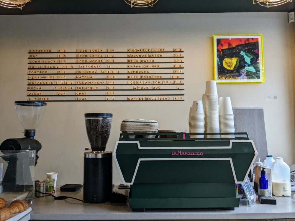 Inside view of Cable Co Coffee in Notting Hill, with La Marzocco espresso machine and grinders on counter, and price board on wall in the background