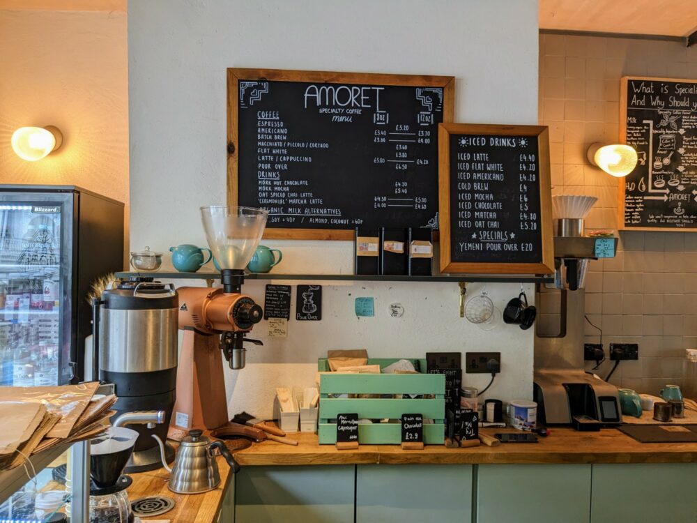Blackboard with prices and coffee options at Amoret Coffee, with a grinder and other coffee equipment on the counter