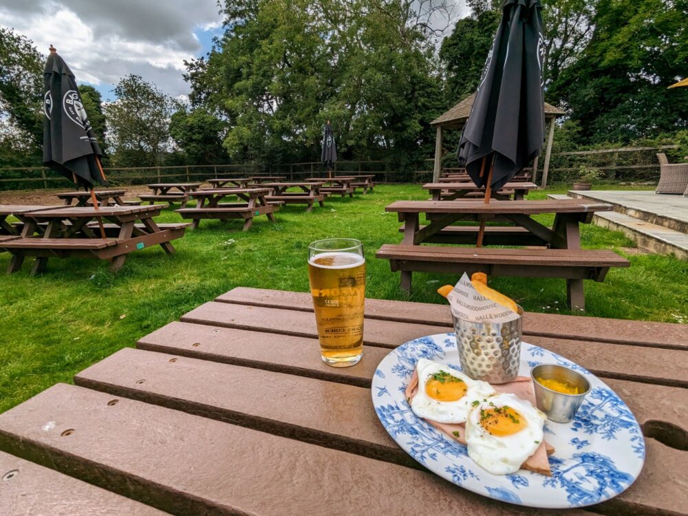 Two fried eggs on slices of ham, with a small basket of chips and a side of piccalilli, on a plate that's sitting on an outdoor table in a pub beer garden. Pint of cider alongside, with several empty outdoor tables in background.