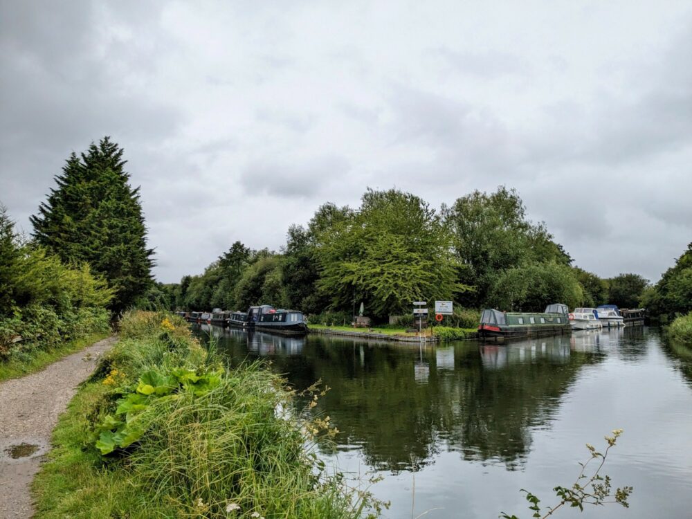 Houseboats moored along the banks of a river under cloudy skies, with a walking path along the left-hand side.