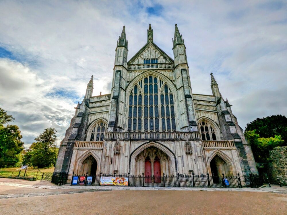 Front of Winchester Cathedral, with large entranceway and stained glass windows visible. Grassy area visible alongside to the left. Blue sky with white clouds above