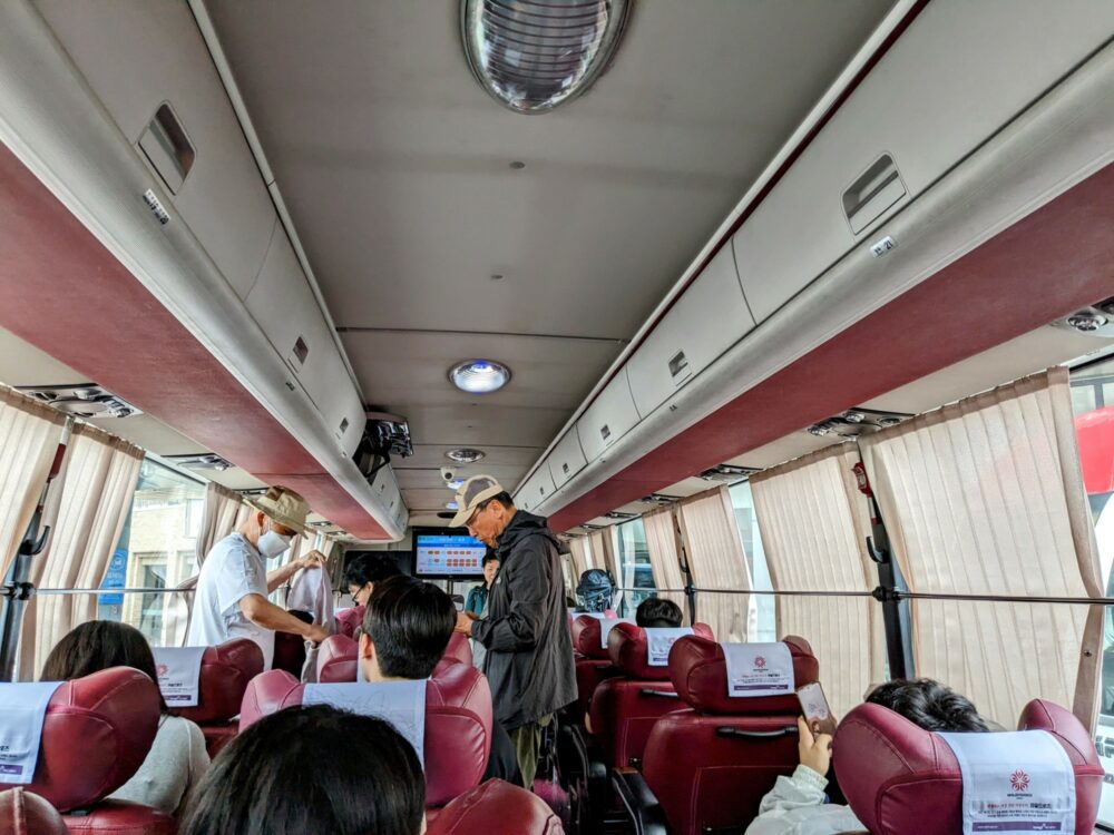 Interior of an "excellent" grade bus in South Korea. Each row has three red leather seats, two on the left and one on the right. Most seats are occupied by seated passengers, with two people standing. Small overhead compartments visible on both sides of the bus. Windows mostly covered by curtains.