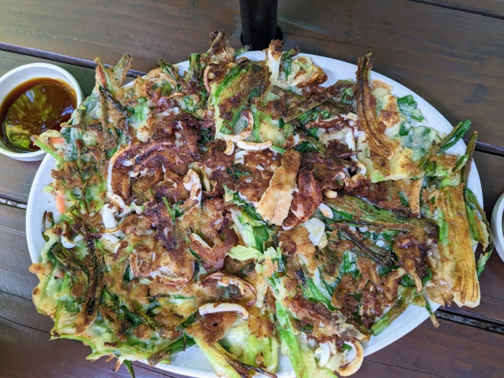 Haemul pajeon (seafood and green onion) pancake on a wooden table, with a small bowl of sauce alongside