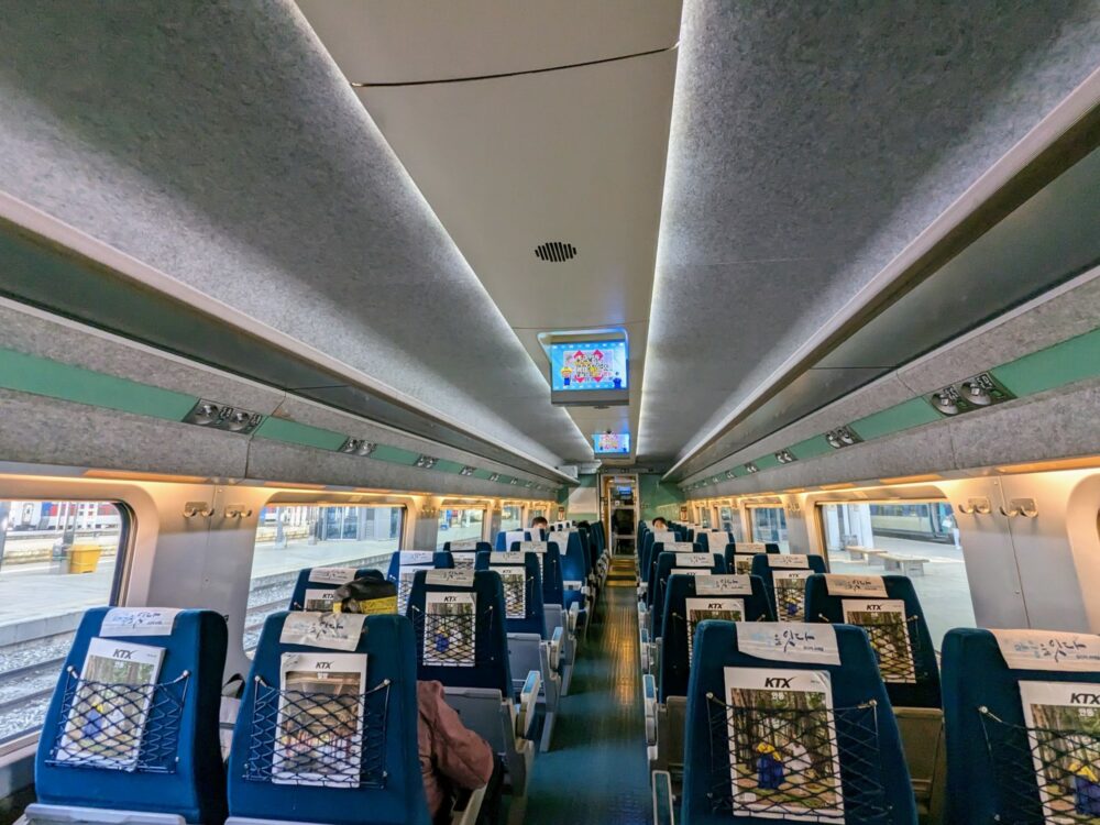 Inside of a KTX high-speed train carriage in South Korea. Many rows of seats, arranged in two by two formation with aisle in the middle. A few seated passengers are visible. A TV screen on the roof is displaying cartoonish characters. Platforms and tracks are visible through the windows.