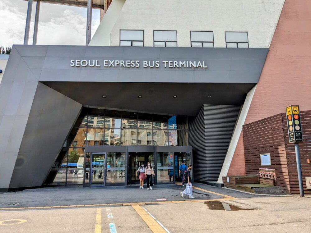Outside view of a bus station in Seoul, with large sign saying "Seoul Express Bus Terminal" above the entrance doors. A few people walking in and out.