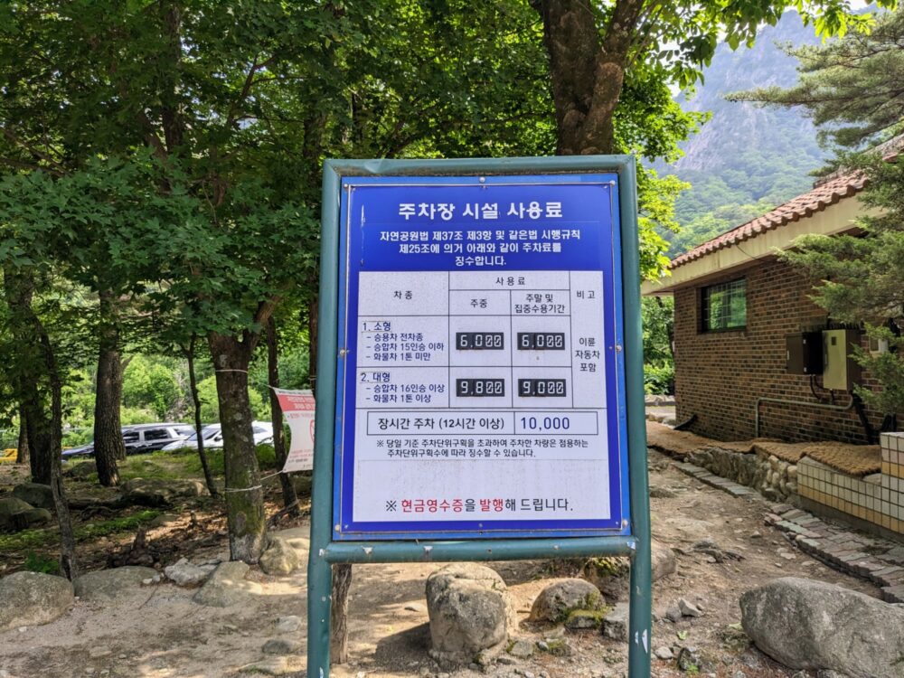 Metal-framed sign (in Korean) showing parking fees at Seoraksan National Park. Rocks on the ground nearby, with trees and cars behind and a brick building to the right. Mountain slopes visible in the background.