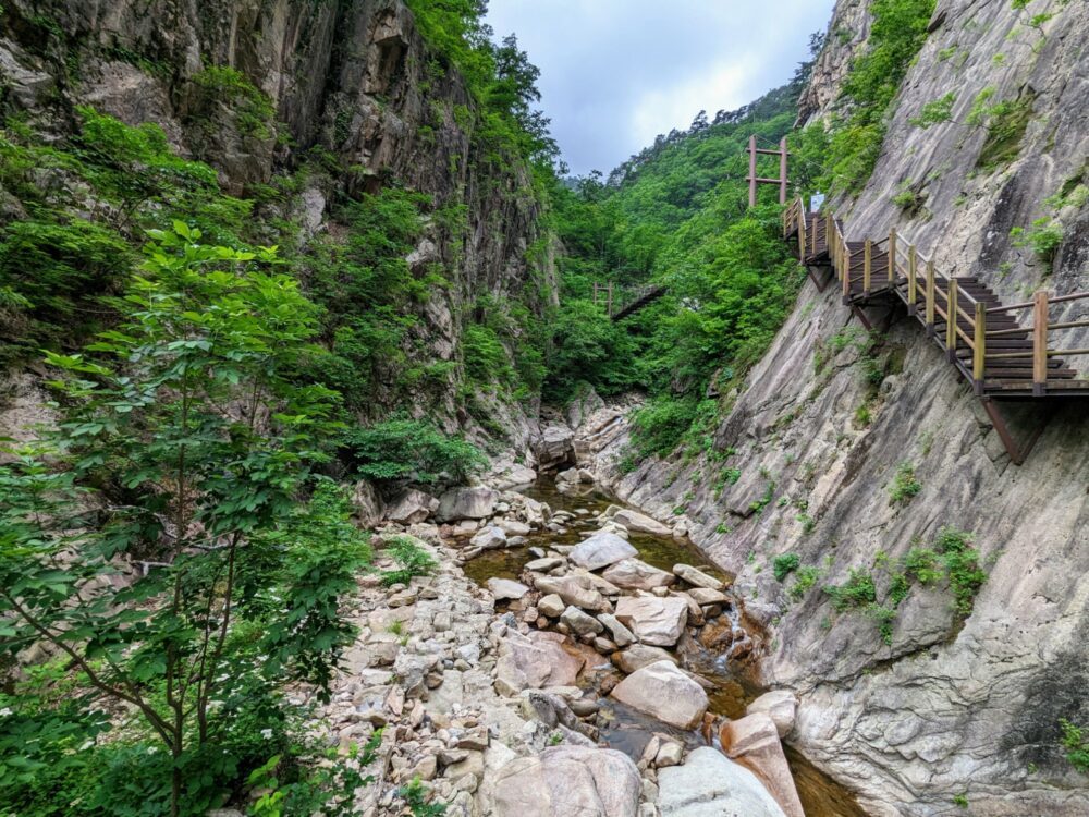 Narrow river gorge with shallow, rocky stream at the bottom. Staircase attached to steep cliff on right-hand side. Suspension bridge visible a little further along the gorge.