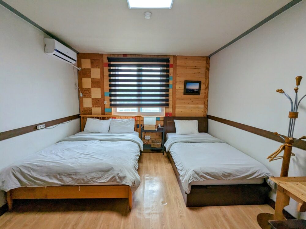 Hotel room with one double and one single bed, with wooden floor and back wall. Window and small painting on back wall, white painted side walls and ceiling, with coat stand and partial view of desk and chair on the right. Air conditioner on the left wall.