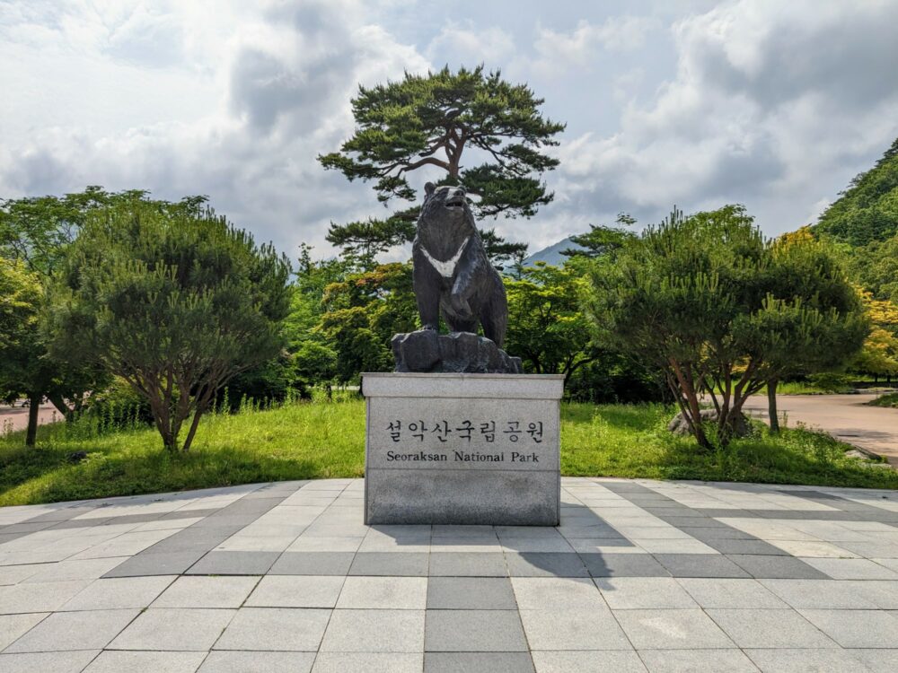 Statue of a bear standing on a concrete plinth with "Seoraksan National Park" written in English and Korean. Concrete paving stones around the statue, with a grassy, tree-filled area behind.