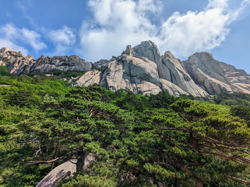 View towards large, rocky, jagged peaks in Seoraksan National Park, South Korea. Dense tree canopy below the mountain range, blue sky with some clouds above