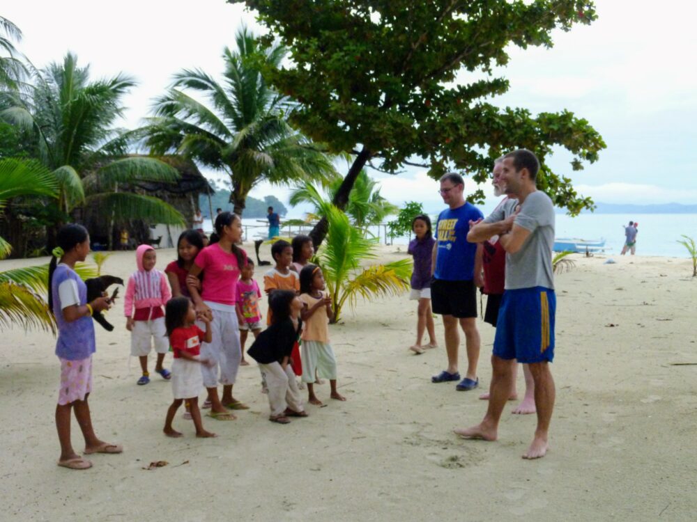 Group of people on a beach: some schoolchildren, some adults. Palm and other trees in the background. Ocean visible behind. Cloudy skies.