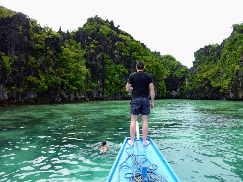 Man standing on front of boat, woman swimming in water in front, limestone cliffs ahead