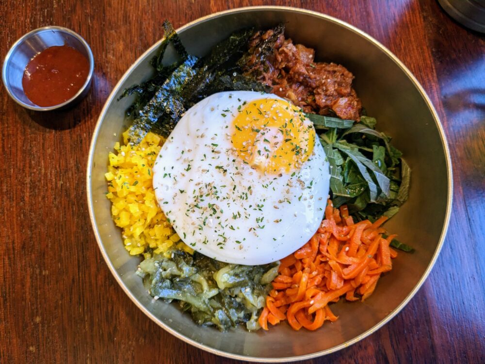 Overhead view of bowl on a wooden table, with a small metal bowl of chili sauce alongside. In the bowl are tuna, rice, carrots, and other vegetables arranged separately, with a fried egg on top.