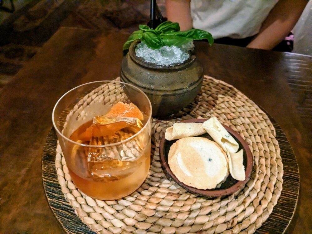 Two cocktails on a woven placemat on a wooden table, with a small plate of crackers alongside. One cocktail is in a clay cup, with shaved ice and decorated with green leaves, the other is in a plain glass with an orange garnish.