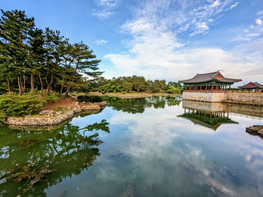Donggung Palace and Wolji pond in Gyeongju. A small lake with no ripples on the surface, reflecting trees on the bank and historic-looking Korean buildings in the background.