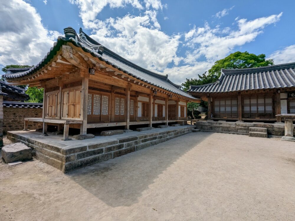 Two wooden houses in a traditional Korean style, sitting on tiled stone platforms around a courtyard.
