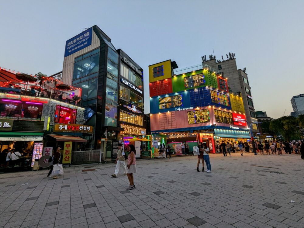 Street scene at sunset in Hongdae, Seoul, with brightly-lit buildings alongside a pedestrianized street. One building appears to be made from colorful shipping containers, with 'M Playground' on the side