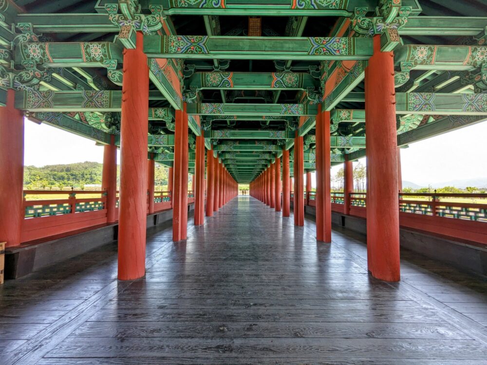 View along the length of a long wooden bridge, with red pillars and green painted beams that are helping to support a second level above. Hills are visible in the distance.