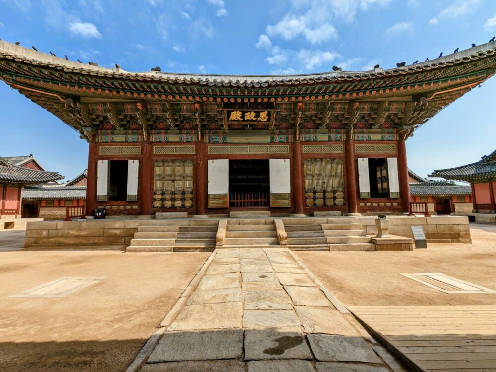 Ornate building in traditional Korean style at Gyeongbokgung Palace in Seoul, with other similar buildings partially visible on both sides. A stone path runs up to the front of the building.