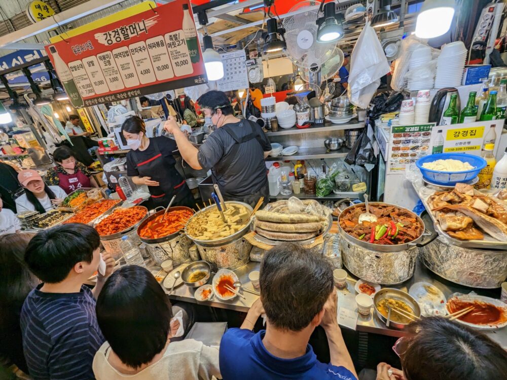A busy food stand at Gwangjang market in Seoul, South Korea, with several bowls and plates of different food on the counter, staff cooking and preparing food behind, and people sitting in front eating.