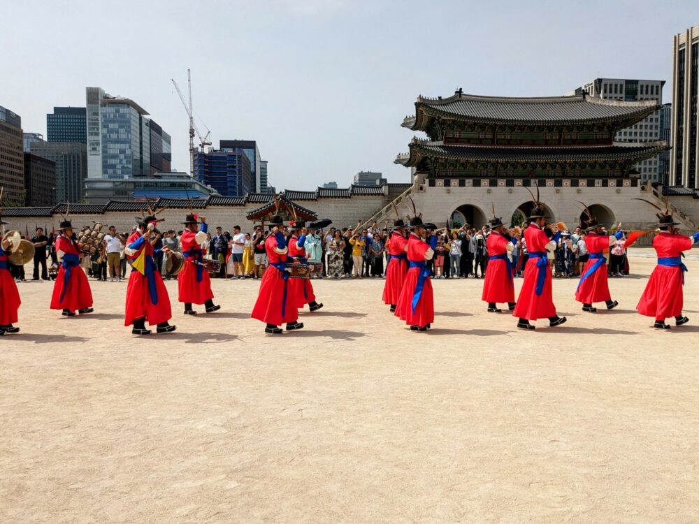 Several men with ceremonial dress and weapons marching in a sandy courtyard inside a palace in Seoul, South Korea, with a crowd of people behind