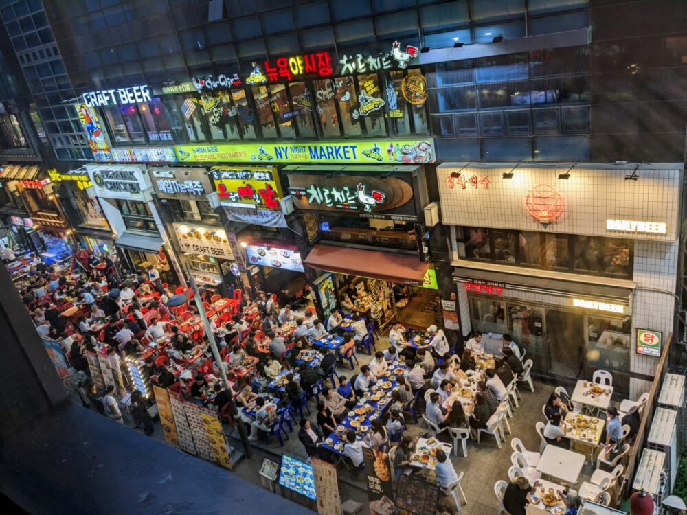 Many chicken and beer restaurants alongside each other on a small street in Myeongdong, Seoul, with outside tables that are almost all occupied by diners. Price boards and neon signs advertise each restaurant's offerings.