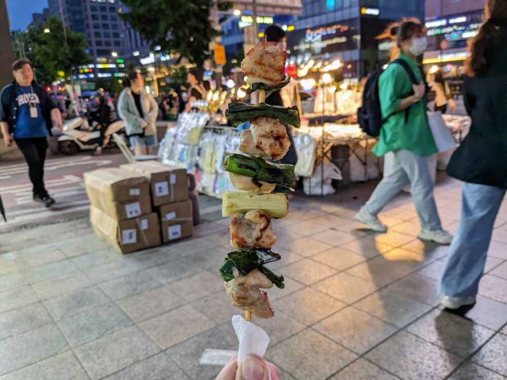 Closeup of chicken skewer being held in hand at a night market in Hongdae, Seoul, with bright street scene slightly blurred in the background.