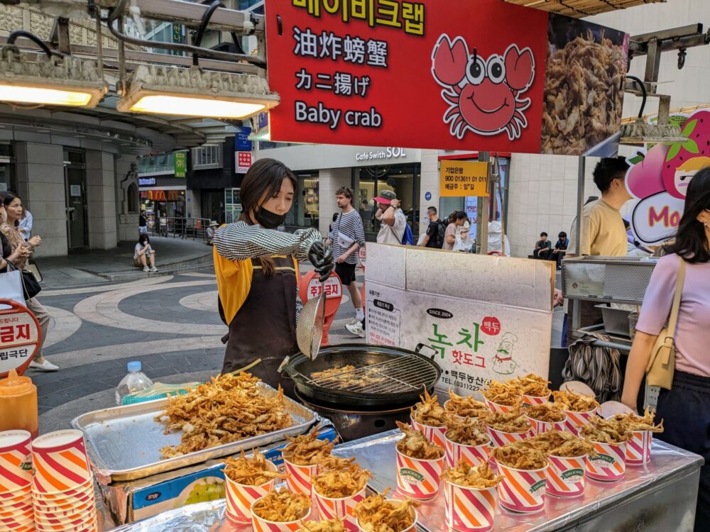 Several containers of deep-fried baby crab at a street food stand, with a woman cooking more crab on a BBQ grill behind.