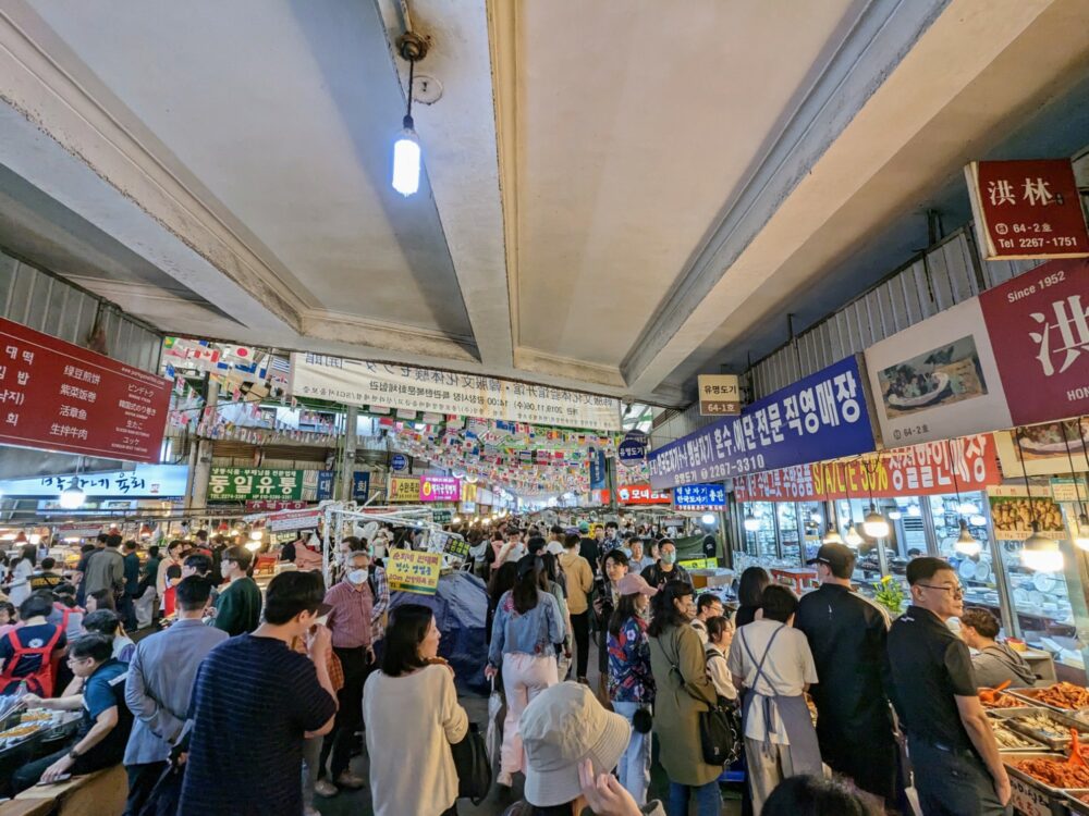 Crowds of people at an indoor market in Korea, with many food stands visible and dozens of signs advertising what's available to purchase.
