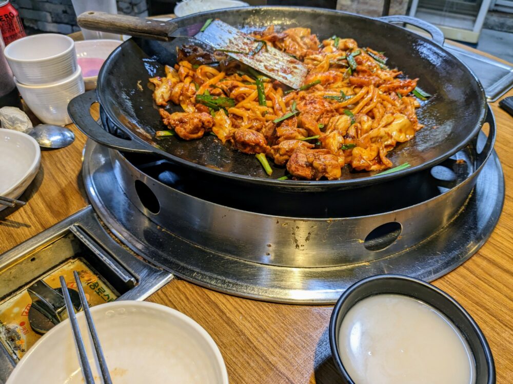Large iron pan on a gas cooker inset into a wooden table, with a stir-fried chicken dish inside and a metal spatula sitting on top. Various bowls and cups are visible alongside on the table.