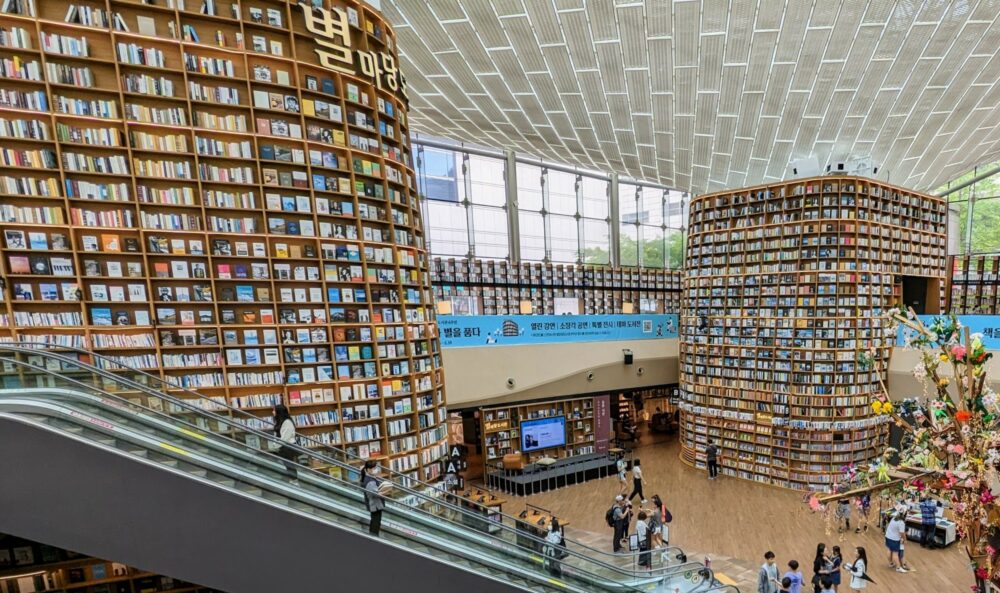 View of the Starfield Library inside COEX Mall in Seoul. Two huge two-storey bookcases full of books stretch from floor to ceiling, with escalators beside one of the bookcases. A fake tree on the ground floor is partially visible.