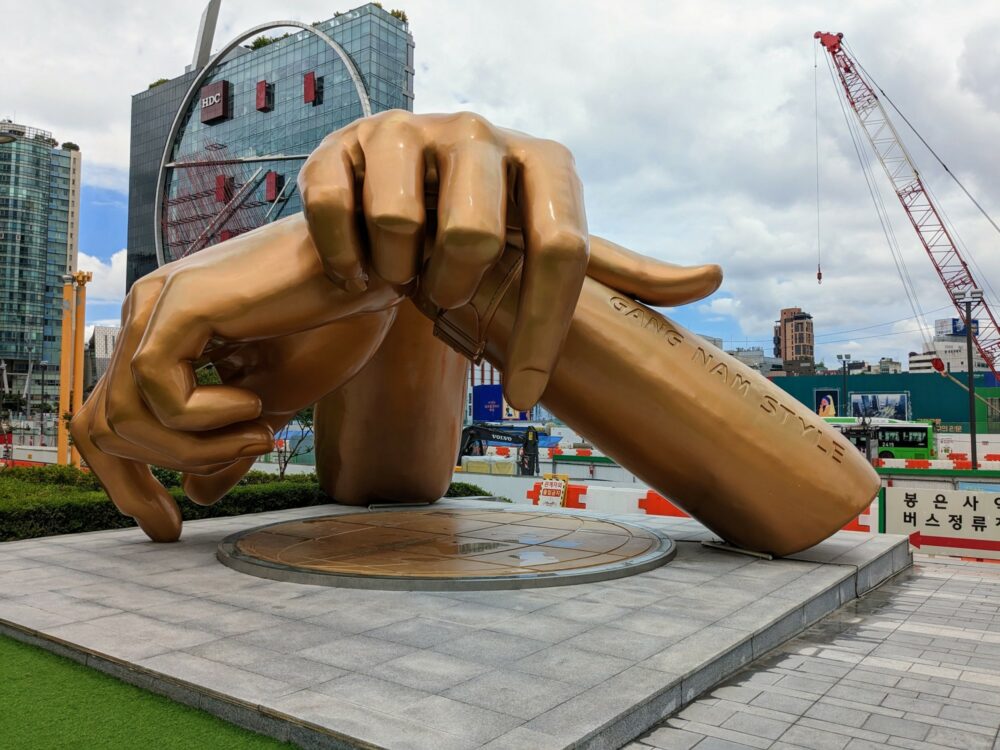Large golden sculpture of two hands folded over top of each other, with "Gang Nam Style" engraved on one of the forearms. Tall office buildings and a construction crane are visible in the background.