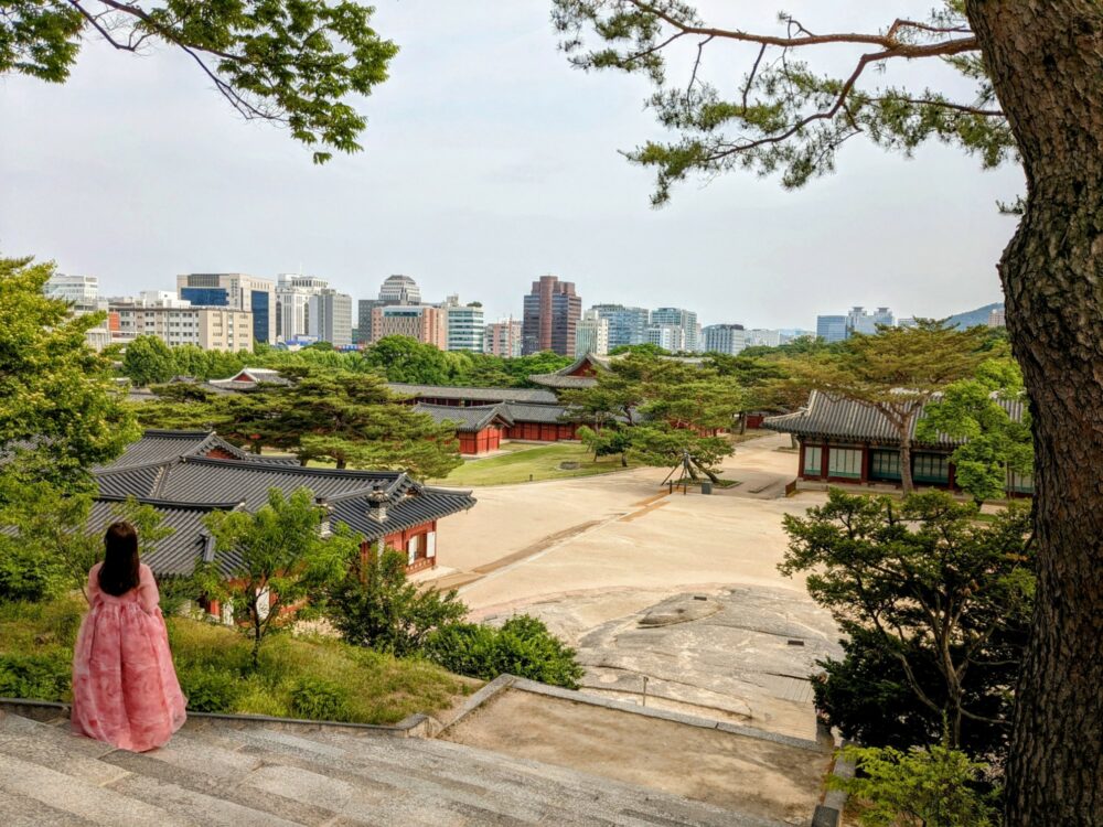 Woman in traditional Korean dress standing on stone steps that lead down to a large courtyard with ornate buildings surrounding it. Trees line the stairs and courtyard. Skyscrapers visible in the distance.