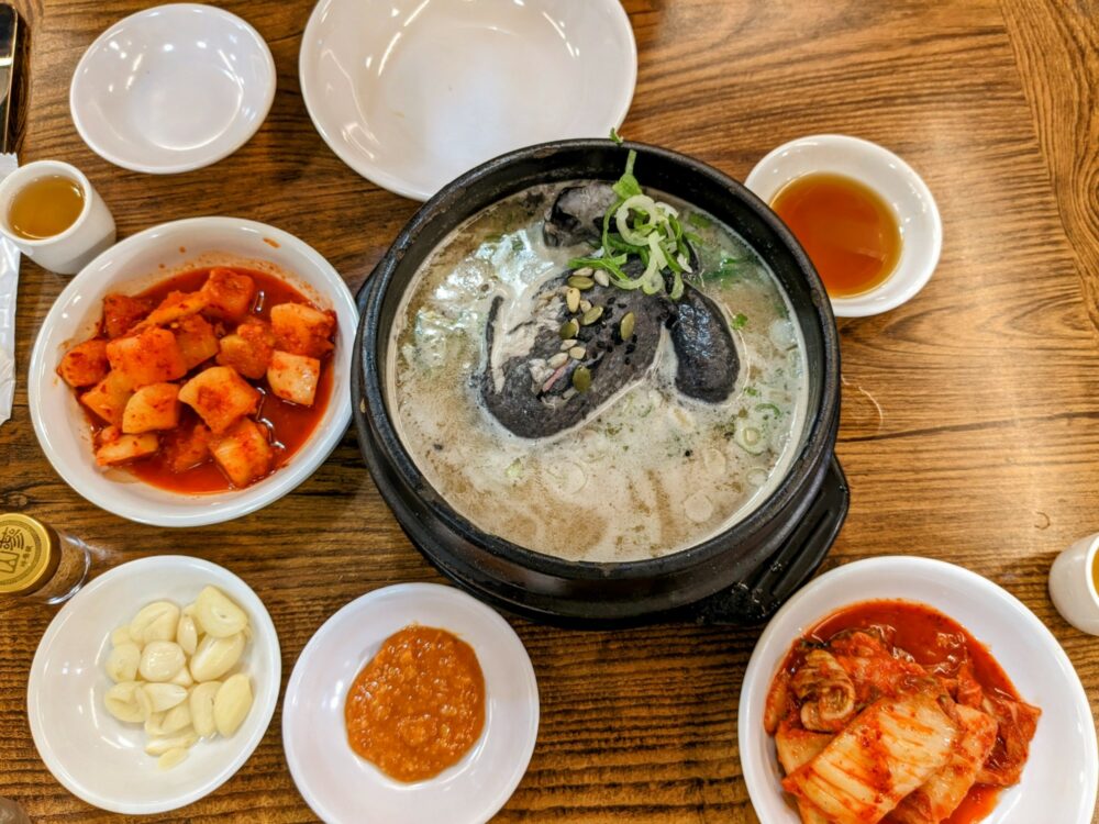 Large bowl of soul with black chicken in, and several dishes of kimchi, garlic, and dipping sauces alongside.