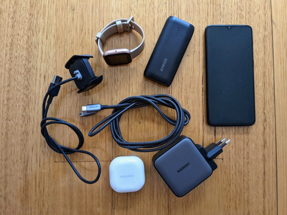 Smartphone, power bank, smartwatch, wall charger, two charging cables, and wireless earbuds case on a wooden floor