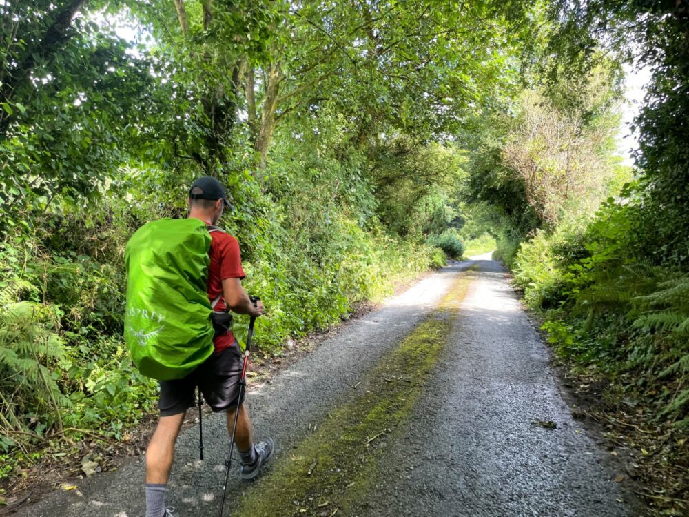 View from behind and to the side of a man hiking with a backpack covered by a pack cover down a country lane