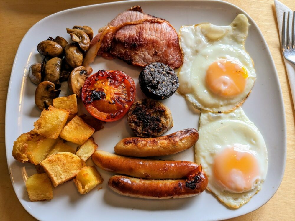 Large breakfast on a white plate, with fried eggs, sausages, fried potatoes, mushrooms, bacon, black and white pudding, and grilled tomato.