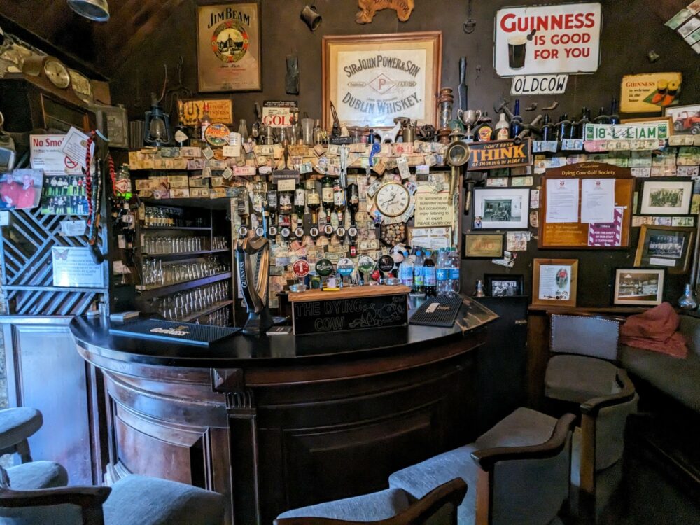 Interior of a small, quirky pub in Ireland with a few chairs around a circular bar. Many notices, bottles, photos, and other memorabilia are behind the bar